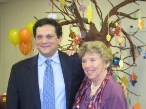 The late Dr. Ted Rubenstein, left, and Marilyn Richman, right. They have their arms around each other, smiling, and are dressed in formal clothing. Behind them are yellow and orange balloons and a Papier-mâché tree with hand-made ornaments hanging from the branches.