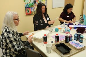 3 women sit at an art table with paints, shaving cream, and paper.