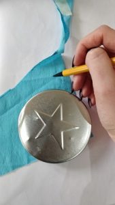 A person uses a metal disc to draw a curved edge onto a piece of blue fabric.