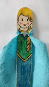 A popsicle stick drawn to look like Queen Elsa from Frozen with a small blue cape fastened with an elastic band around the neck.