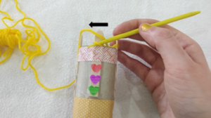 Yellow yarn wrapped around 4 pegs of a knitting doll, one loop is being pulled over another on the same peg.