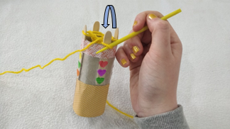 A loop of yellow yarn being pulled over another loop on a peg of a handmade french knitting doll.