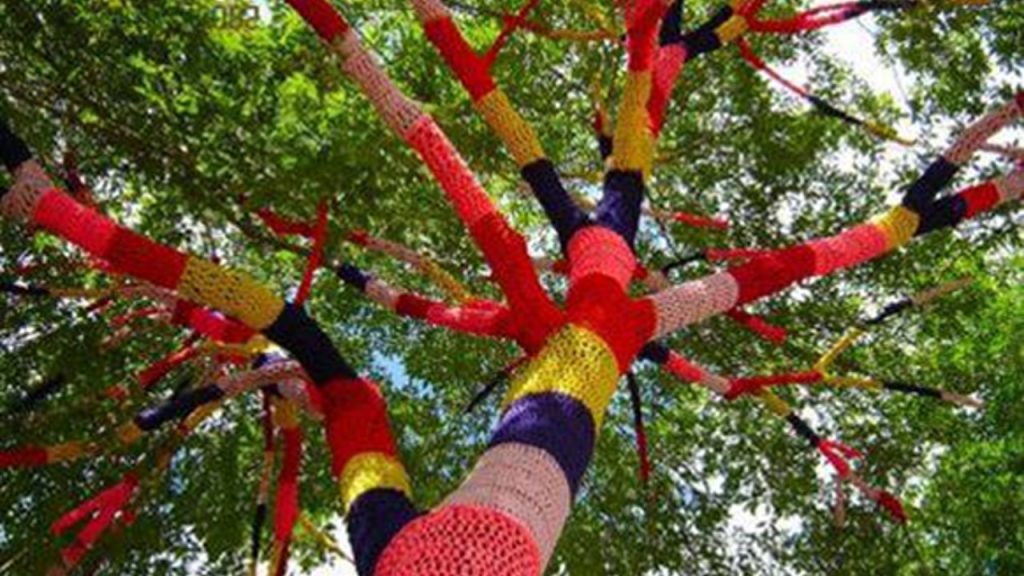 A green tree with its trunk and branches covered by colorful knitted yarn